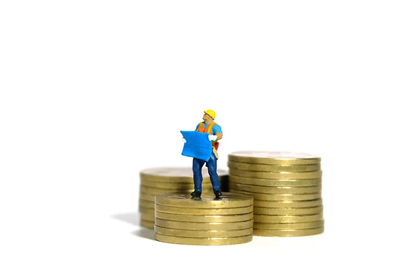 Eine kleine Bauarbeiterfigur steht auf gestapelten Münzen und hält einen Bauplan in der Hand. Symbolisiert Investitionen in die Entwicklung. Helles und fokussiertes Bild.